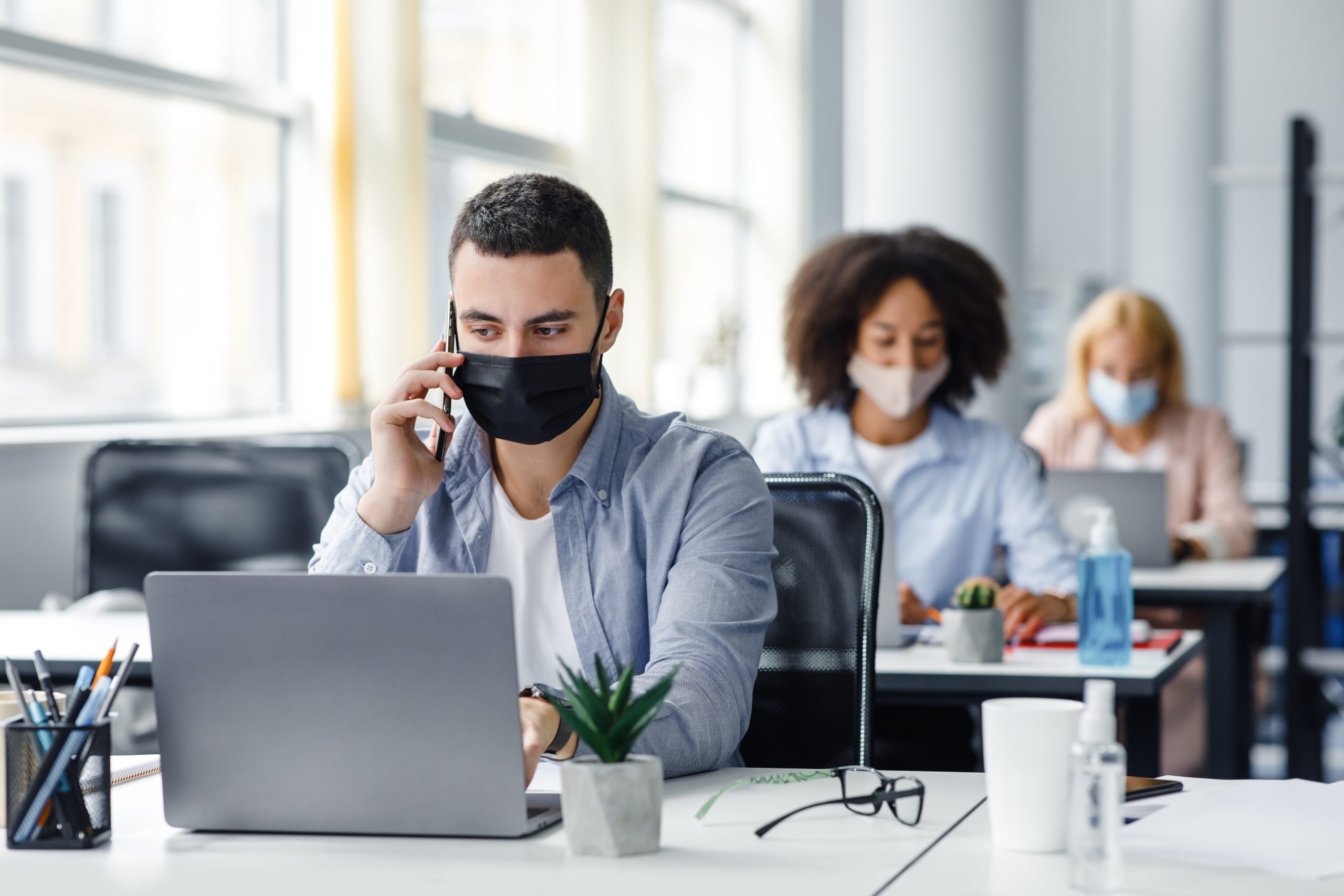 Customer consultation by phone remotely at returning to work after quarantine. Millennial man in protective mask with smartphone looks at laptop at workplace